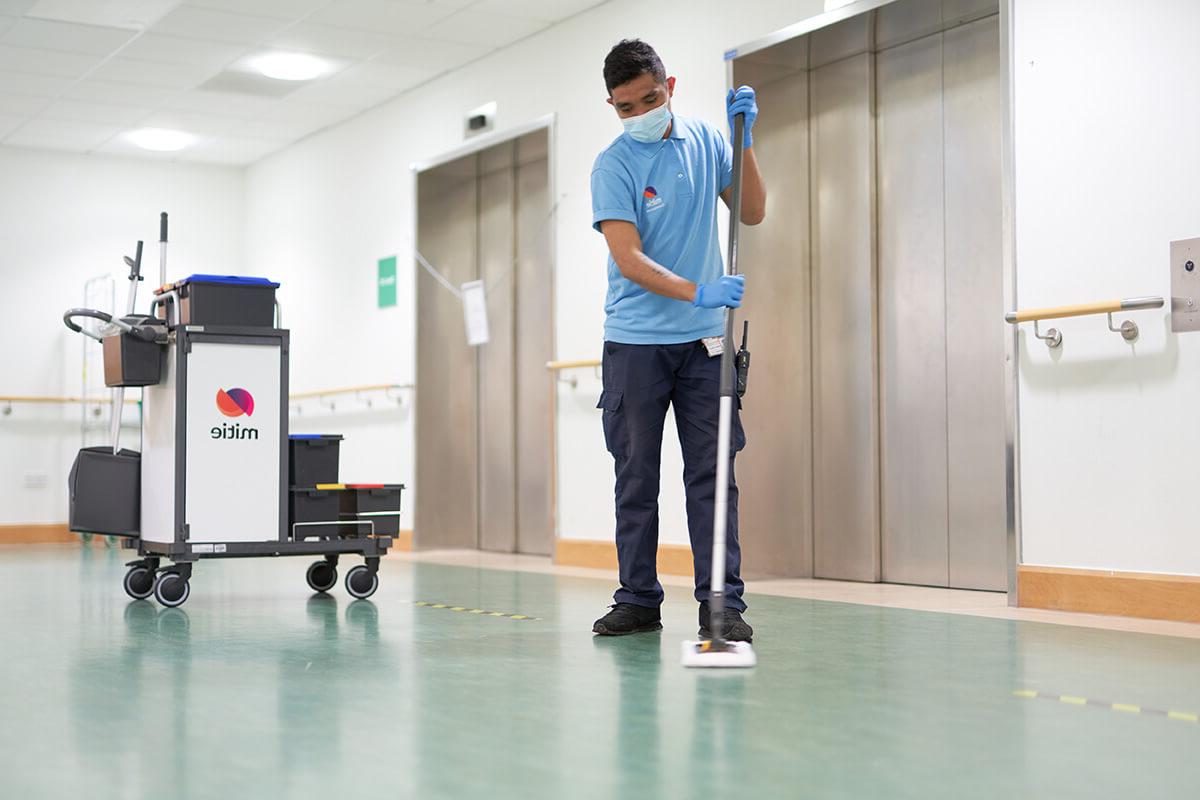 一名米蒂员工在医院走廊用扫帚打扫地板, 背景是一辆米蒂牌清洁车